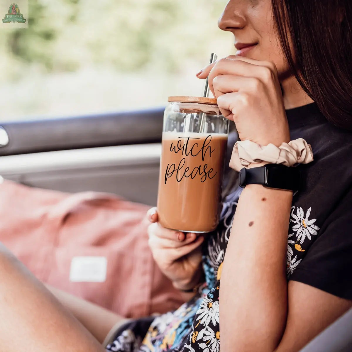 A woman with long brown hair sits in a car, sipping iced coffee from her Witch Please 17oz glass cup with a bamboo lid and stainless steel straw. She wears a floral shirt, scrunchie, and smartwatch.