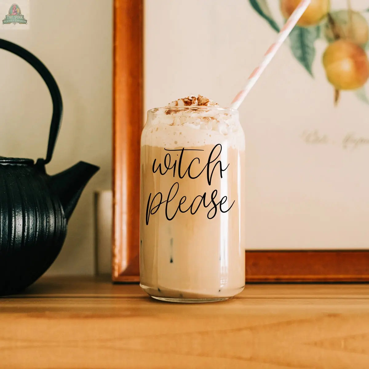 The Witch Please 17oz glass cup is filled with iced coffee, topped with whipped cream and cinnamon, and features "witch please" on the front. A striped stainless steel straw is in the drink, with a black teapot and framed fruit art in the background.