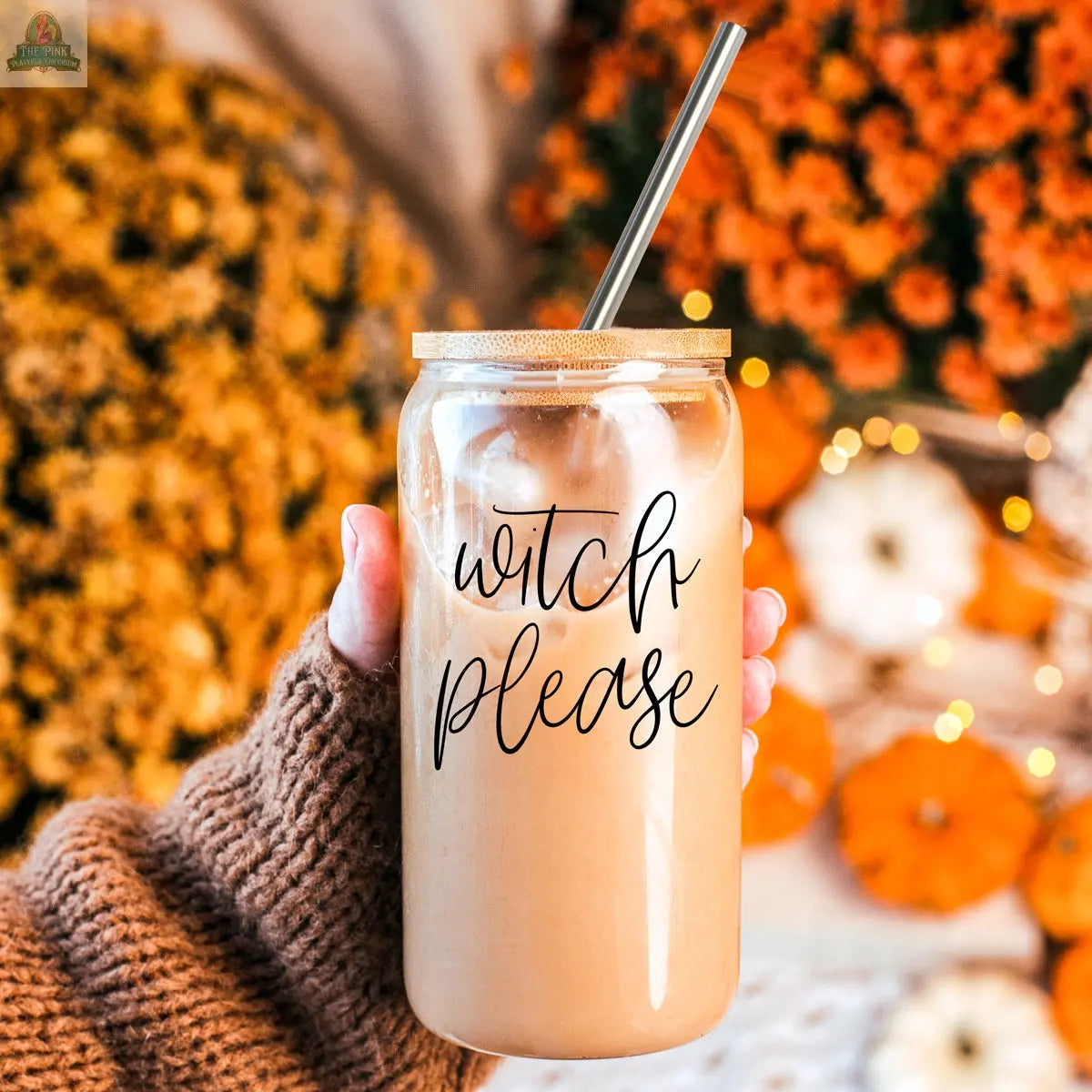 A hand in a brown knit sleeve holds the Witch Please 17oz glass cup with bamboo lid and stainless steel straw. “Witch please” is printed on the glass. Orange and white pumpkins and autumn flowers appear in the background.