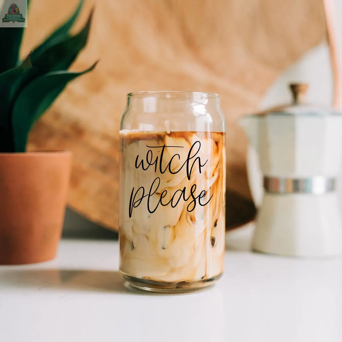 The Witch Please 17oz glass cup, filled with iced coffee and cream, sits on a white surface; a potted plant, moka pot, and bamboo lid with stainless steel straws are in the background.