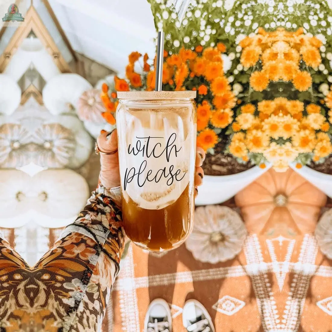 A hand holds the Witch Please 17oz glass cup with a bamboo lid and stainless steel straw. The cup is filled with iced coffee, while orange flowers, white pumpkins, and a fall-themed patterned rug accent the background.