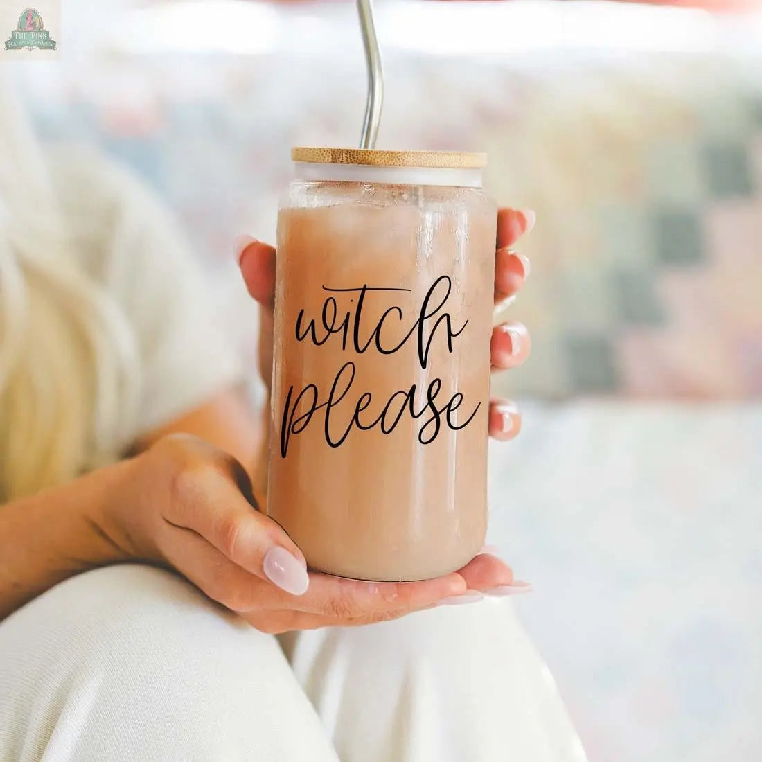 A person holds the Witch Please 17oz glass cup filled with iced coffee, topped with a bamboo lid and stainless steel straw. The cursive "witch please" inscription stands out against a softly blurred backdrop.