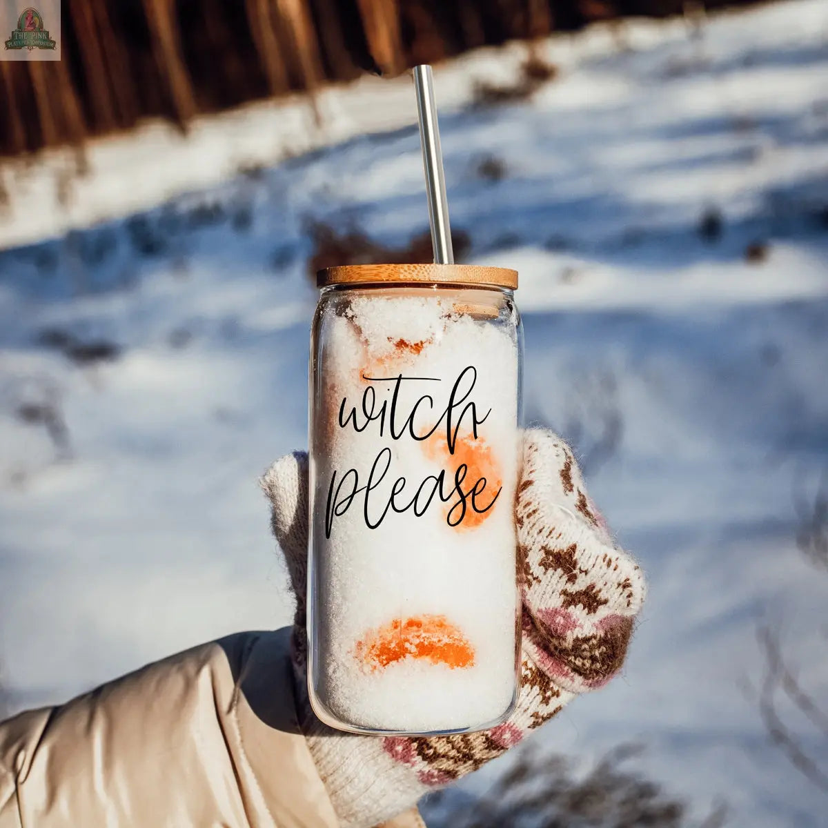 Someone wearing patterned mittens holds the Witch Please 17oz jar, featuring a bamboo lid and metal straw, filled with snow and orange slices. The scene is set against a snowy outdoor backdrop.