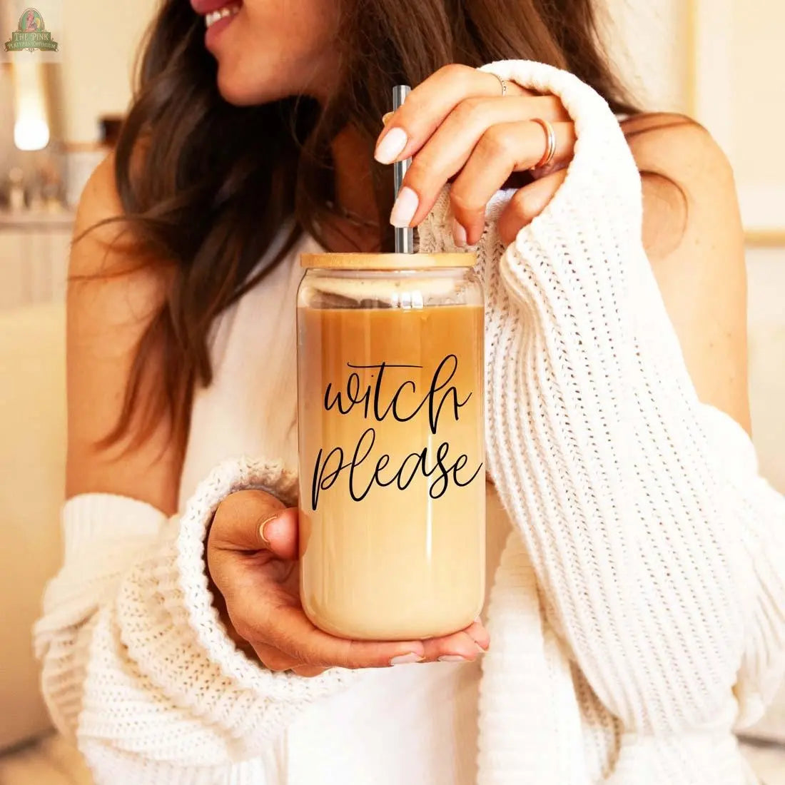 A woman with wavy brown hair holds the Witch Please 17oz glass cup, featuring a bamboo lid and stainless steel straw. She wears a white sweater and ring, smiling slightly as she enjoys her iced coffee.