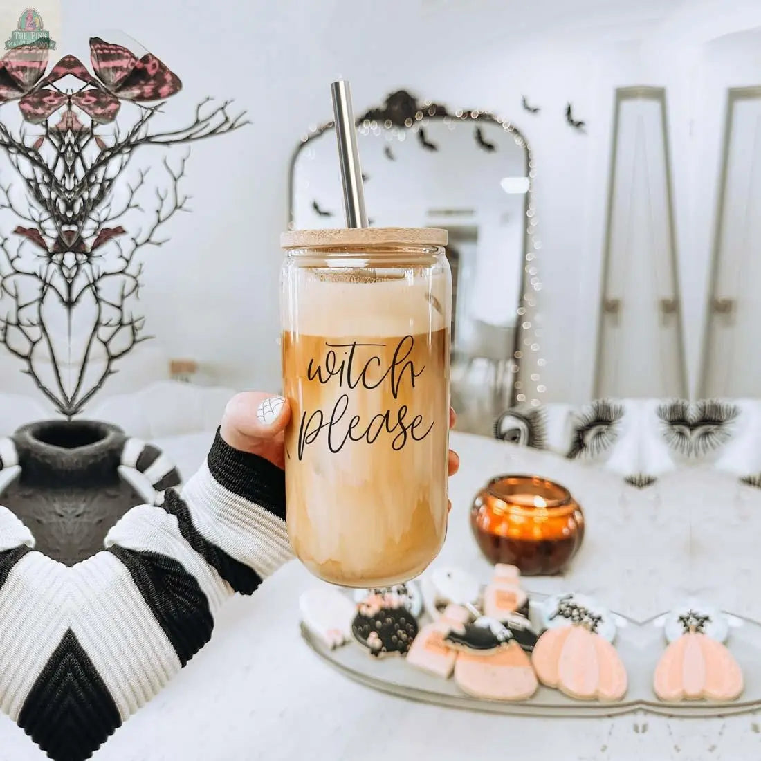 A hand holds the Witch Please 17oz glass cup with bamboo lid and stainless steel straw. Halloween decorations, pumpkin and bat cookies, a lit candle, and a mirror appear in the background.