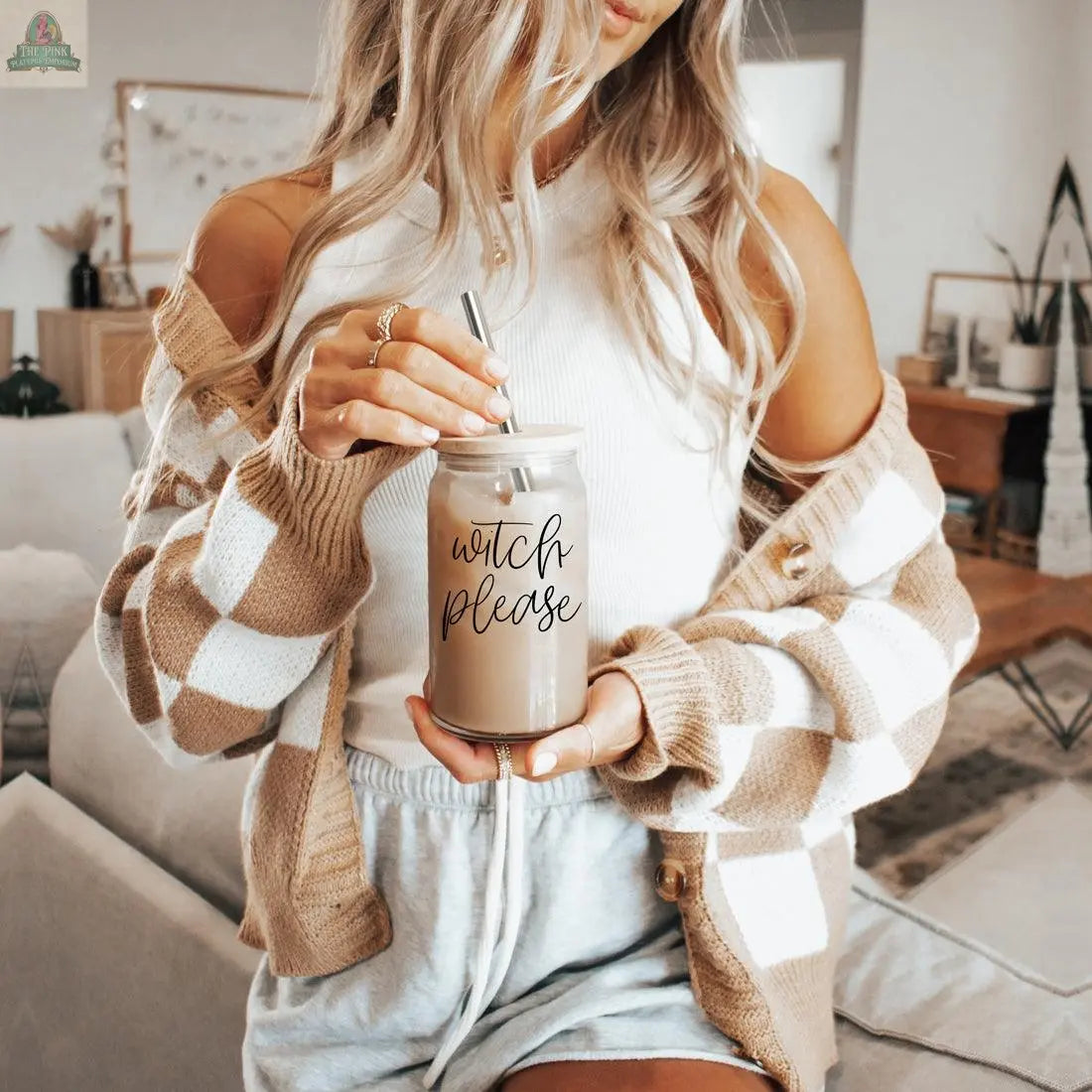 A woman with long blonde hair holds the Witch Please 17oz glass cup with bamboo lid and stainless steel straw in a cozy, sunlit living room. She wears a white top, checkered cardigan, and gray shorts.