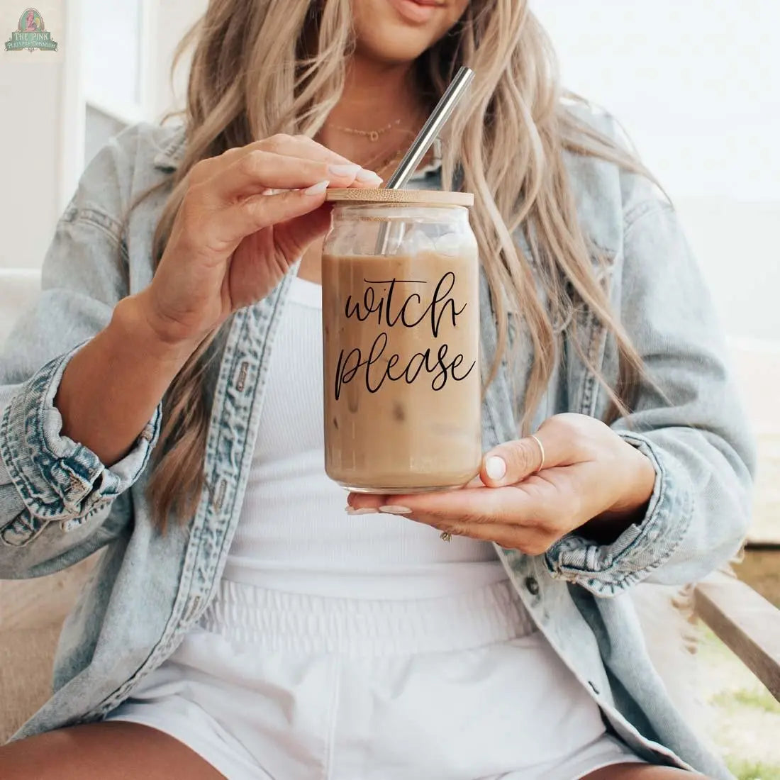 A woman in a denim jacket and white outfit holds the Witch Please 17oz glass with iced coffee. Topped with a bamboo lid and stainless steel straw, it features black "witch please" text as she stirs her drink.