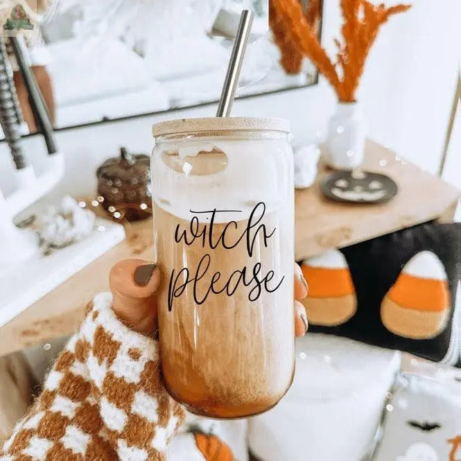 A hand in a checkered sweater holds the Witch Please 17oz glass, filled with iced coffee and cream, topped with a bamboo lid and stainless steel straw. Autumn and Halloween decor, including candy corn pillows, are in the background.