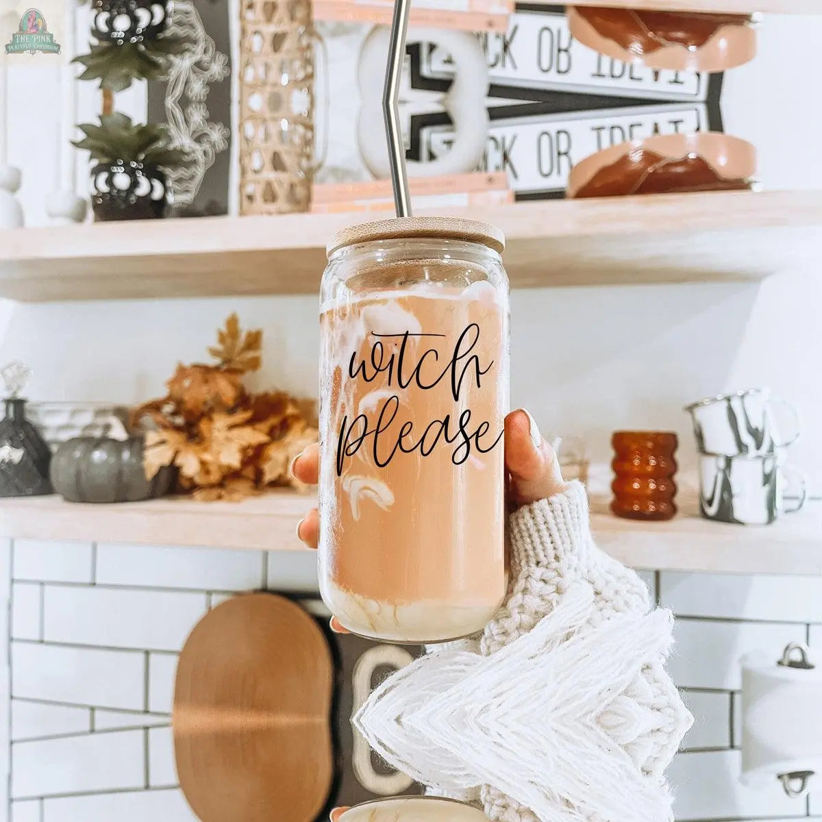 A hand in a cozy sweater holds the Witch Please 17oz glass jar, topped with a bamboo lid and stainless steel straw. Fall decorations, mugs, and shelves can be seen in the background.