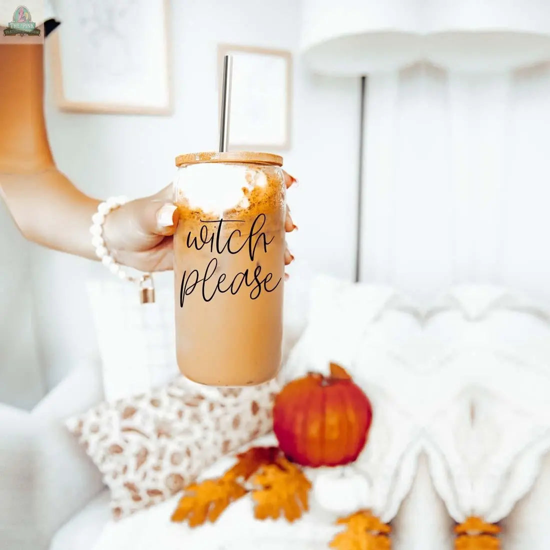 A hand holds an iced coffee in the 17oz Witch Please glass cup with a stainless steel straw. In the background, autumn decorations like a pumpkin, leaves, and a cozy blanket on a couch create a warm, seasonal vibe.