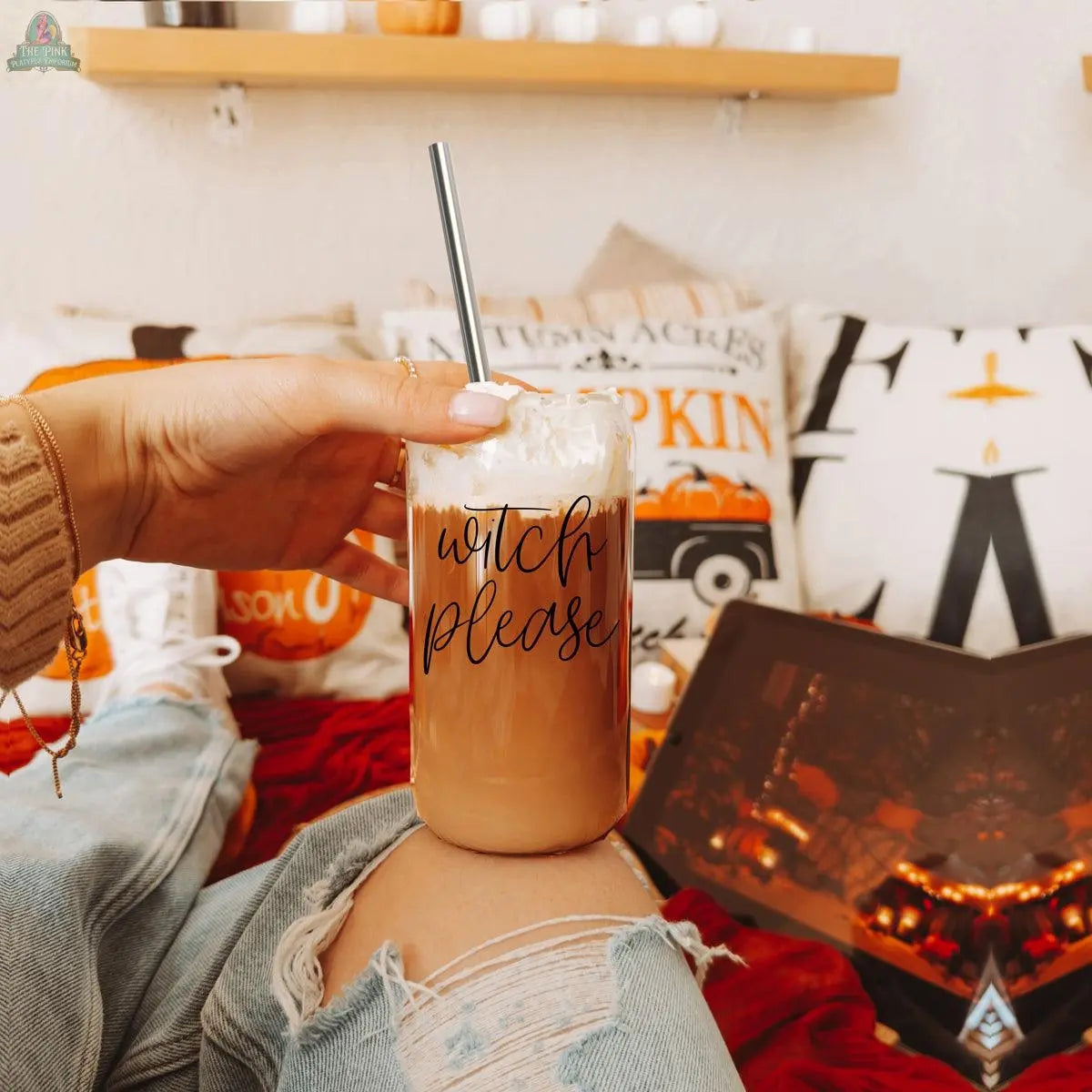 A person in ripped jeans holds a Witch Please 17oz glass cup of iced coffee with whipped cream and a stainless steel straw. Cozy fall pillows and a fireplace decorate the background.
