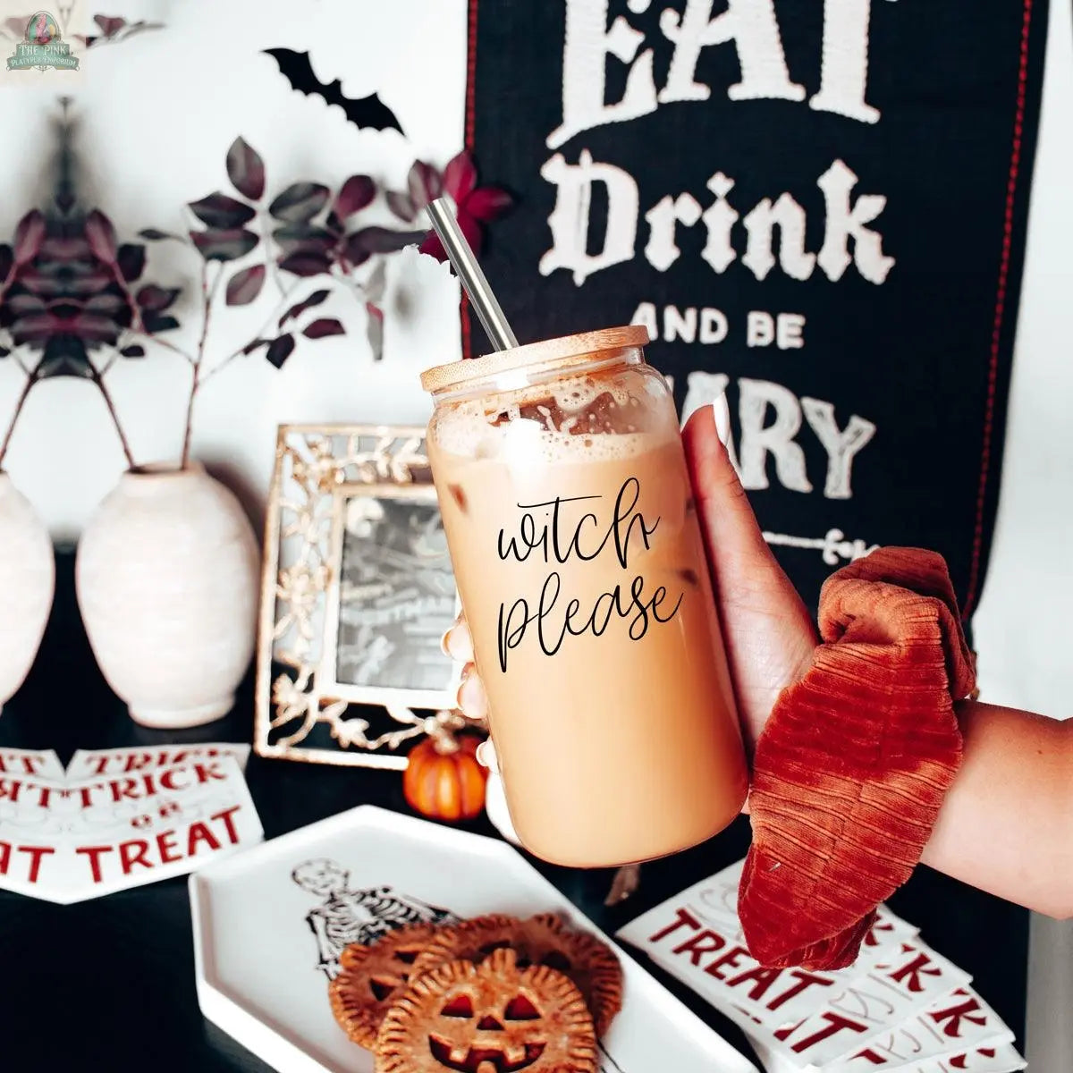 A hand holds the Witch Please 17oz glass jar with iced coffee, bamboo lid, and stainless steel straw; Halloween decorations, mini pies, and trick or treat plates appear in the background.