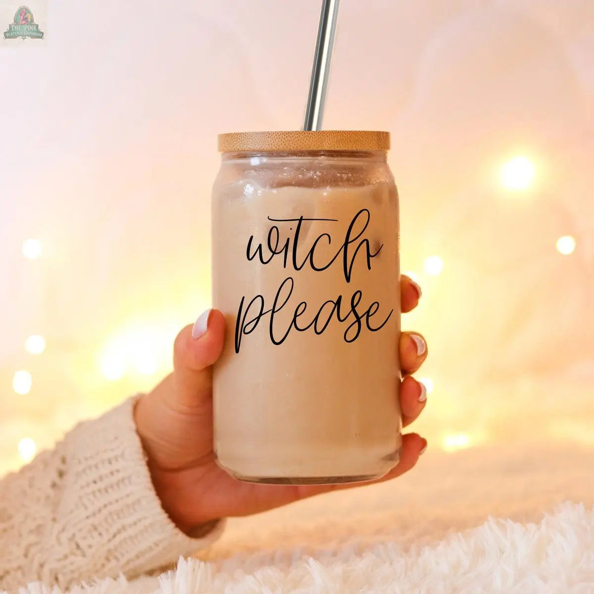 A hand holds the Witch Please 17oz glass cup filled with iced coffee and a stainless steel straw. Cozy fairy lights softly blur in the background, highlighting the cup's cursive “witch please” design.