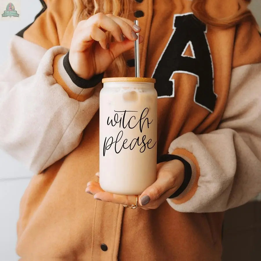 Wearing a tan and black varsity jacket with a large A, someone holds the Witch Please 17oz glass cup, featuring "witch please" in black script, bamboo lid, and straw—adding charm to their stylish iced coffee moment.