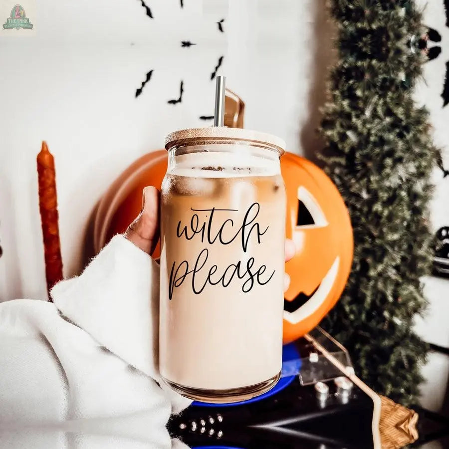 A hand holds a Witch Please 17oz glass cup with a bamboo lid and stainless steel straw, filled with iced coffee, in front of a carved pumpkin, fall decor, and Halloween decorations.