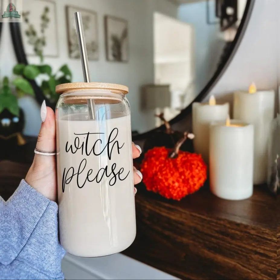 A hand holds the Witch Please 17oz glass cup with a bamboo lid and stainless steel straw. In the background are candles, a red textured pumpkin, a plant, and part of a round mirror on a wooden surface.