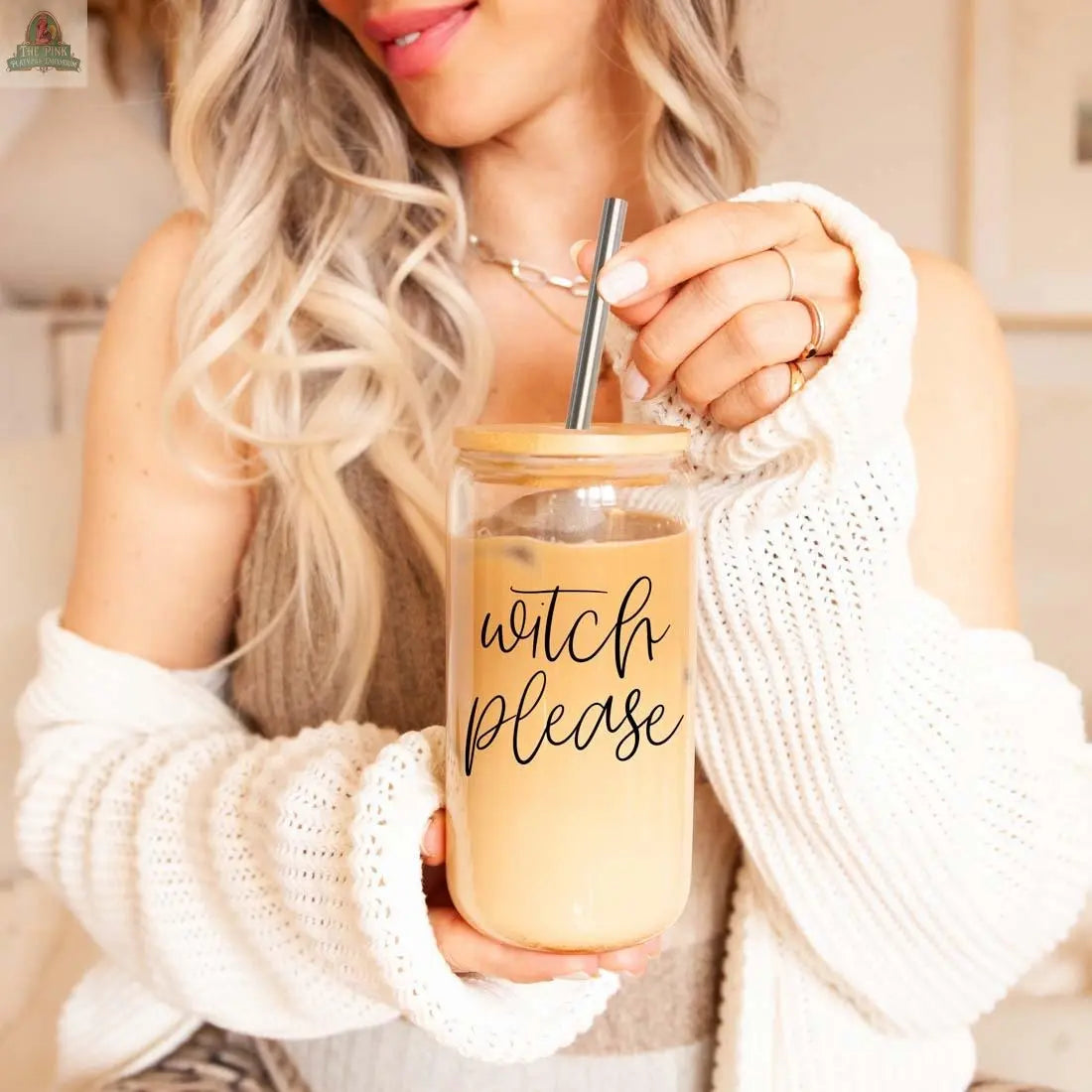 A smiling woman with long blonde hair holds the Witch Please 17oz glass cup with a stainless steel straw. Dressed in a cozy white sweater and layered necklaces, she radiates effortless charm while enjoying her iced coffee.