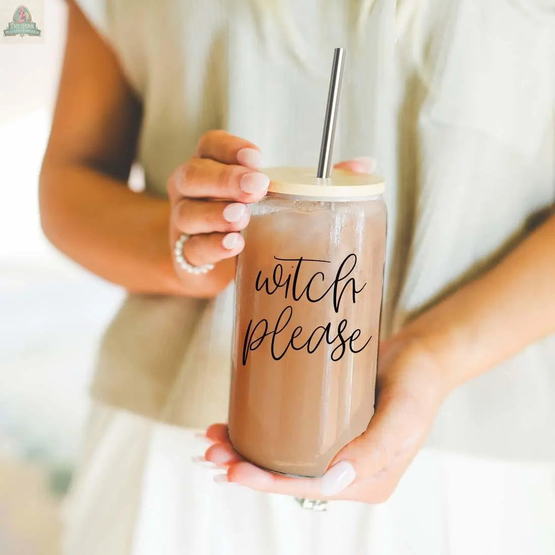 A person with manicured nails holds the Witch Please 17oz glass drinking cup with iced coffee and a stainless steel straw. The stylish jar features "witch please" in cursive. The person wears a light top and a beaded bracelet.