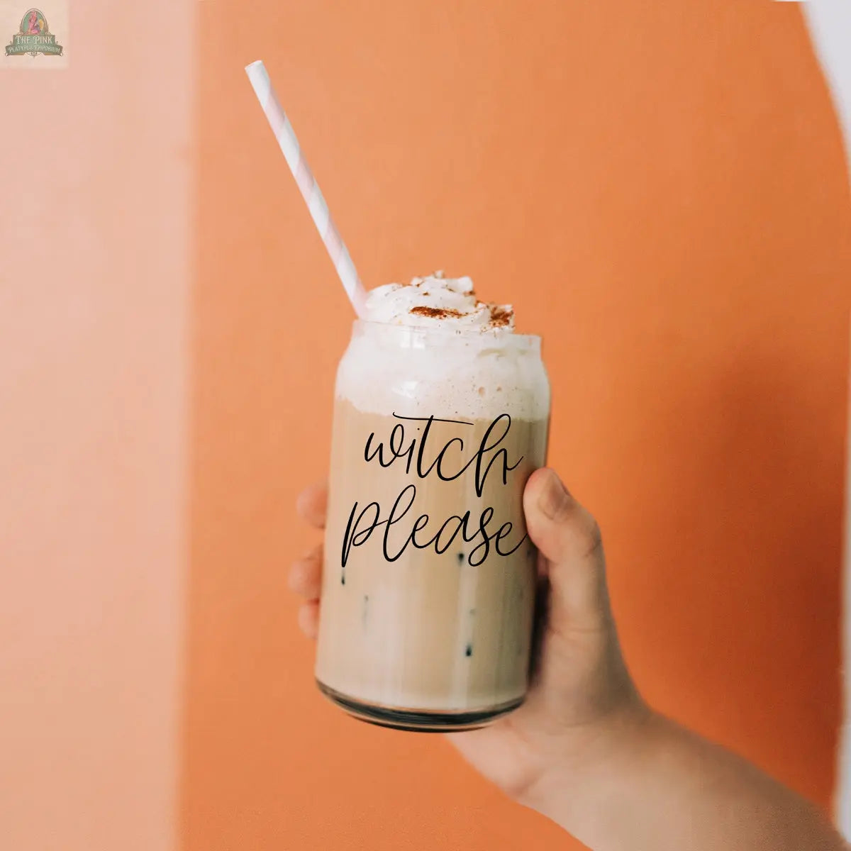 A hand holds a Witch Please 17oz glass filled with iced coffee, topped with whipped cream, cinnamon, and a striped straw. The background is two-tone orange and light beige.