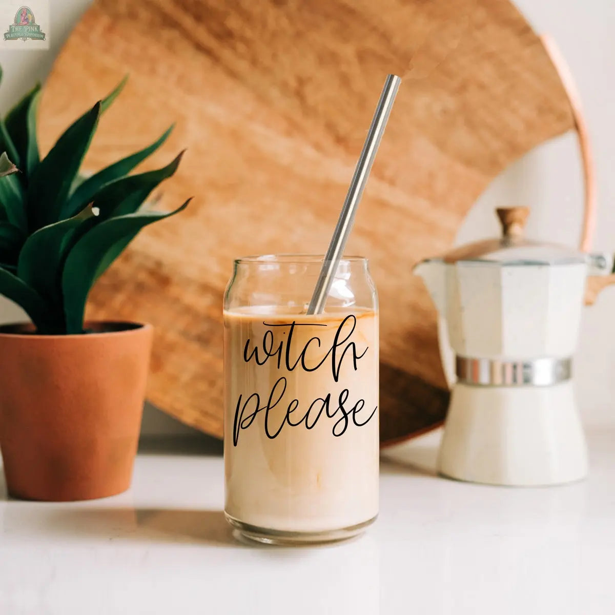 A Witch Please 17oz glass filled with iced coffee and a stainless steel straw sits on a counter. In the background are a plant, a white and silver stovetop espresso maker, and a large wooden tray.