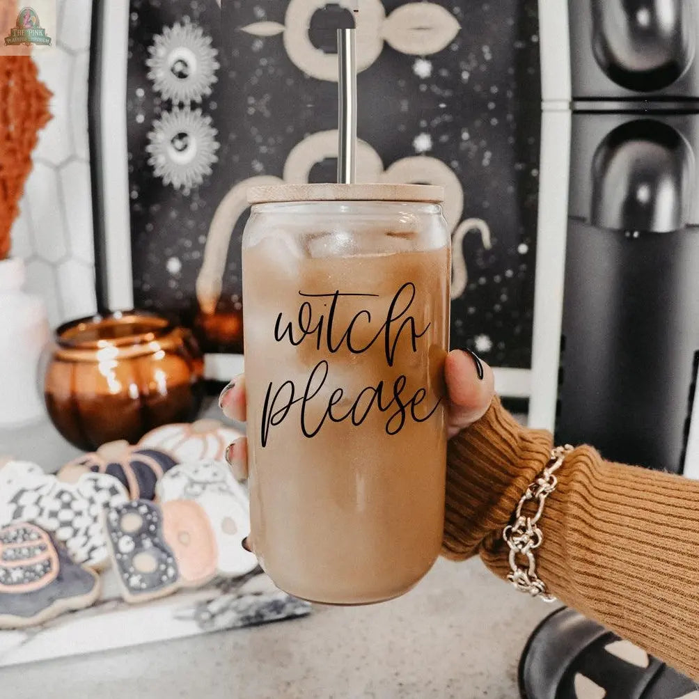 A hand holds the Witch Please 17oz glass cup of iced coffee with a stainless steel straw. The cup displays “witch please.” In the background are decorated cookies, a candle, and mystical decor with celestial patterns.