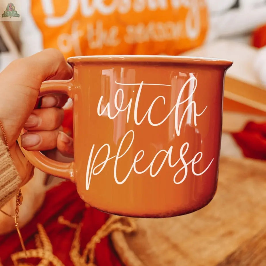 A hand holds the Witch Please Mug, featuring "witch please" in white script. Cozy fall decor, such as an orange pillow and autumn leaves, can be seen in the background.