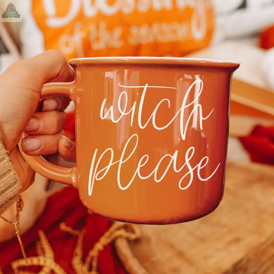 A hand holds the Witch Please Mug, featuring "witch please" in white script. Cozy fall decor, such as an orange pillow and autumn leaves, can be seen in the background.