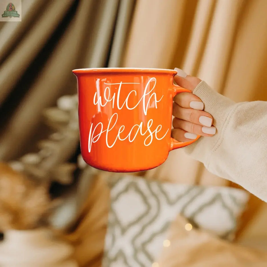 Someone with light-painted nails holds the Witch Please Mug, an orange ceramic cup featuring "witch please" in white script. Cozy, neutral blankets and pillows are visible in the background.