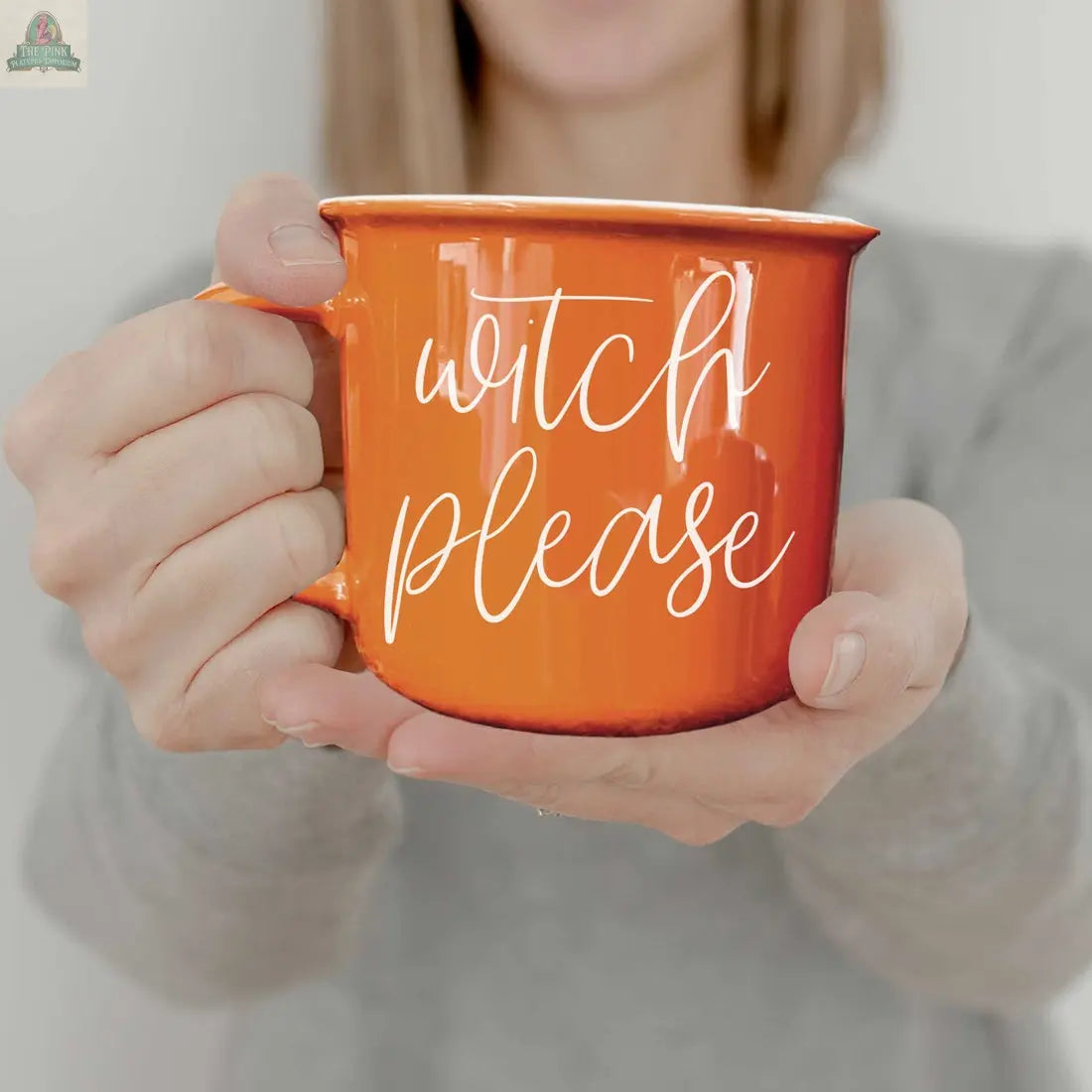 A person in a gray shirt holds the Witch Please Mug, an orange ceramic mug with “witch please” in white script. The playful Halloween coffee mug is in focus, while the person’s face is slightly out of view.