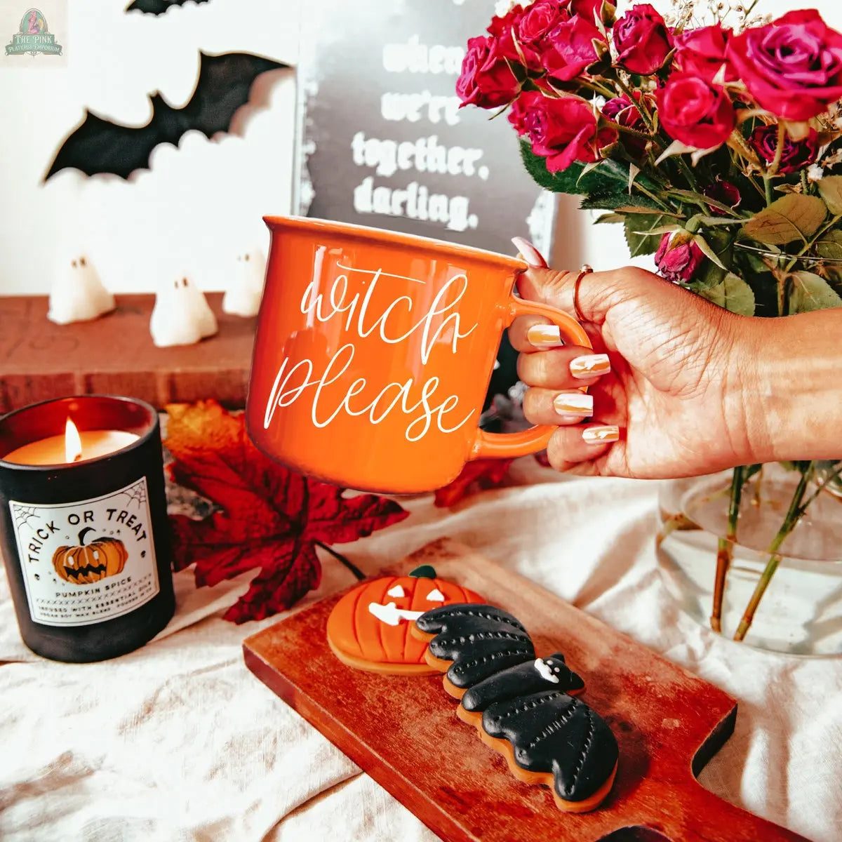 A hand holds the Witch Please Mug, an orange ceramic cup. On the table are Halloween cookies, a Trick or Treat candle, dried roses and leaves, with ghost and bat decorations in the background.