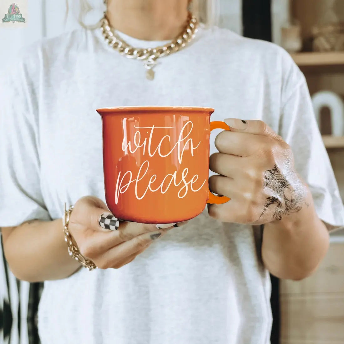 Someone holds the Witch Please Mug—an orange ceramic mug with "witch please" in white script—while wearing a white shirt, gold necklaces, painted nails, and a black ink hand design, creating a stylish Halloween coffee vibe.