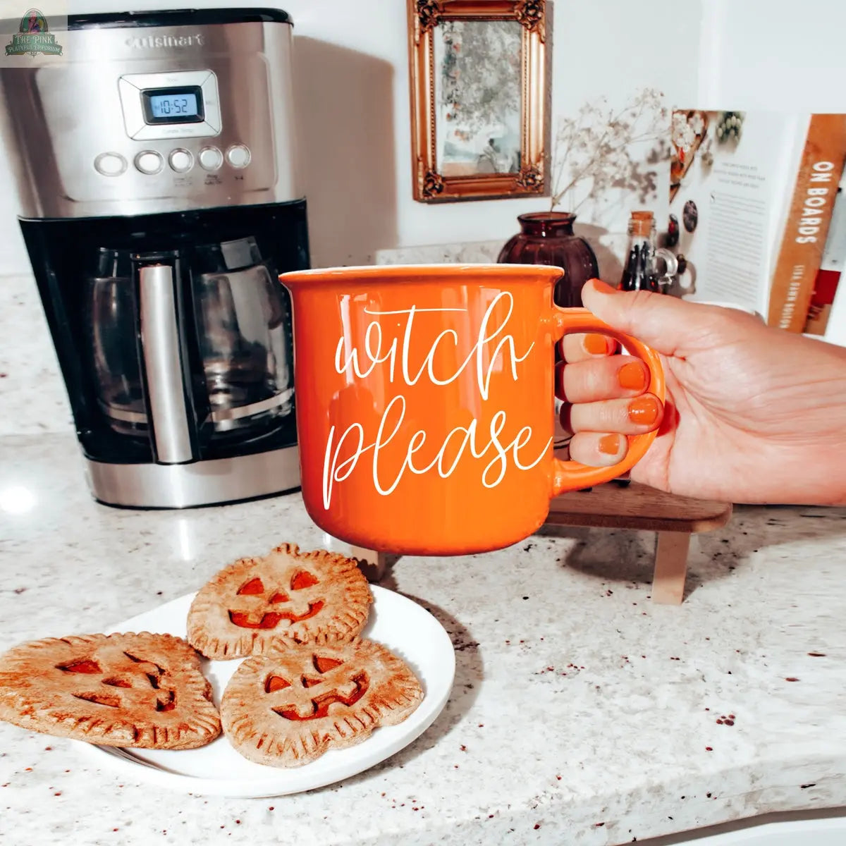 A hand with orange nails holds the Witch Please Mug in a kitchen. On the counter are three pumpkin-shaped pastries, a coffee maker, a mirror, and books.