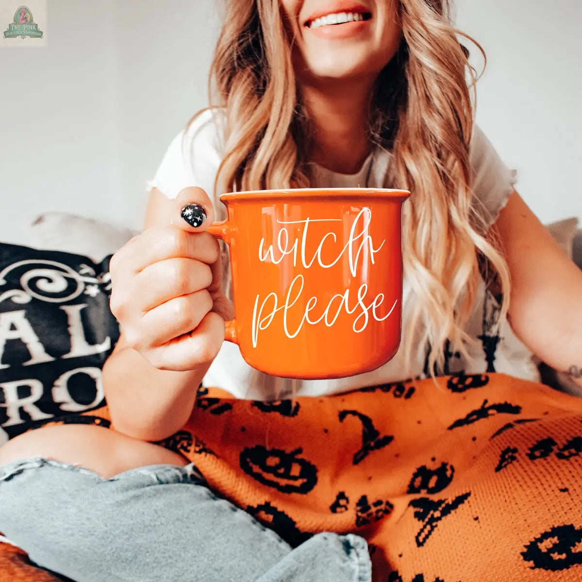 A woman with long wavy hair smiles while holding the Witch Please Mug, an orange ceramic mug. She is sitting on a couch with a Halloween-themed blanket and a decorative pillow in the background.
