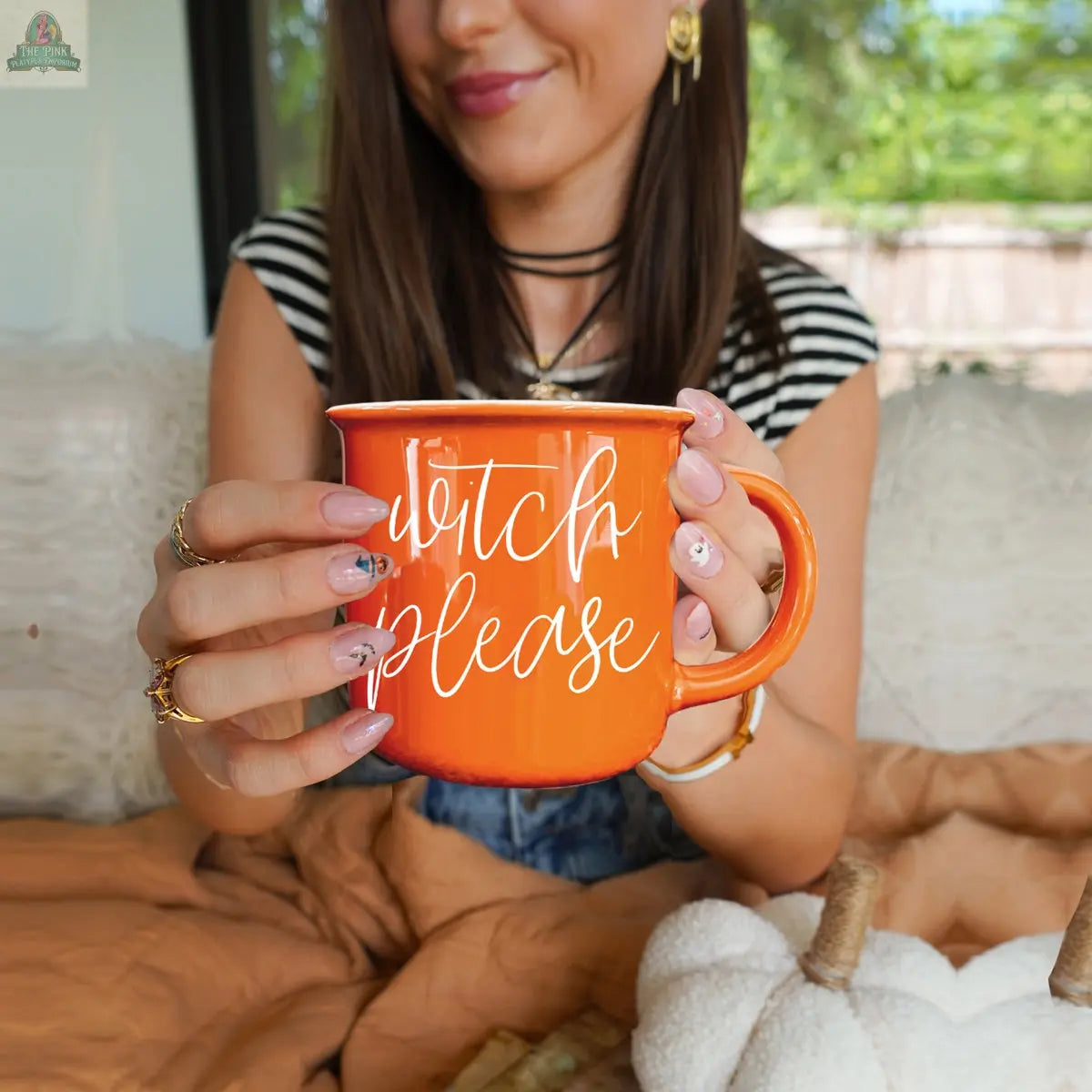 A woman with long brown hair smiles while holding the Witch Please Mug, an orange ceramic mug with white script. She has decorated nails, wears rings, and a white pumpkin decor rests on her lap.
