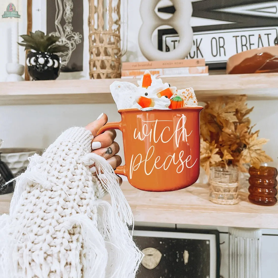 A hand in a cozy, fringed sweater holds the Witch Please Mug, an orange ceramic mug with "WITCH PLEASE" on it, filled with whipped cream and Halloween candy. Autumn decor, plants, and a “Trick or Treat” sign accent the shelves behind.