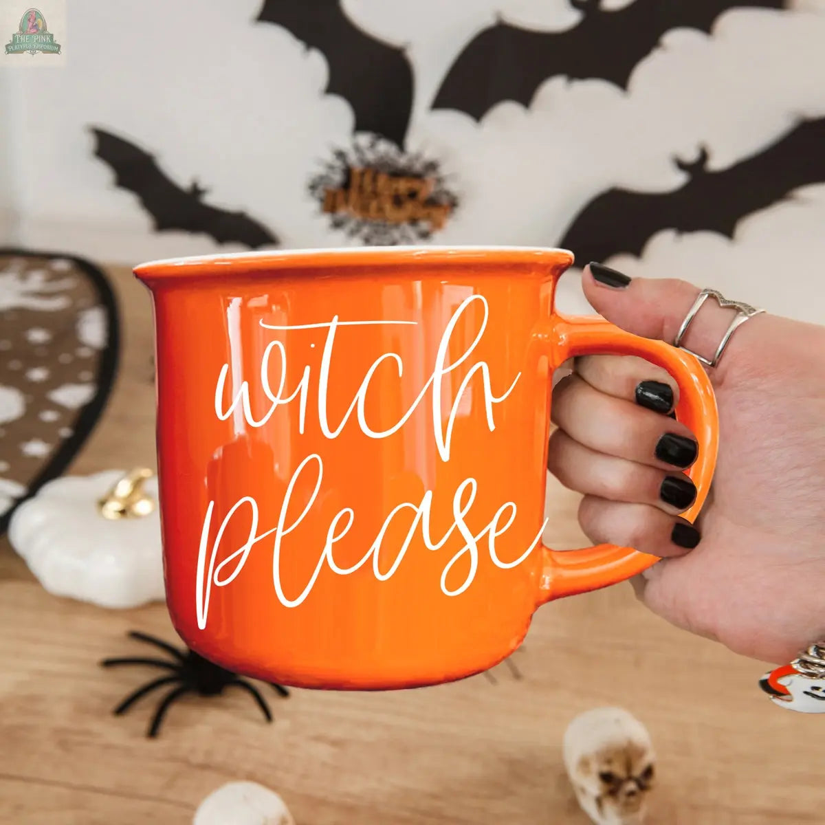 A hand with black nail polish holds the Witch Please Mug, an orange ceramic mug with "witch please" in white script. Halloween decor—black bats, a white pumpkin, and a spider—appear in the background.