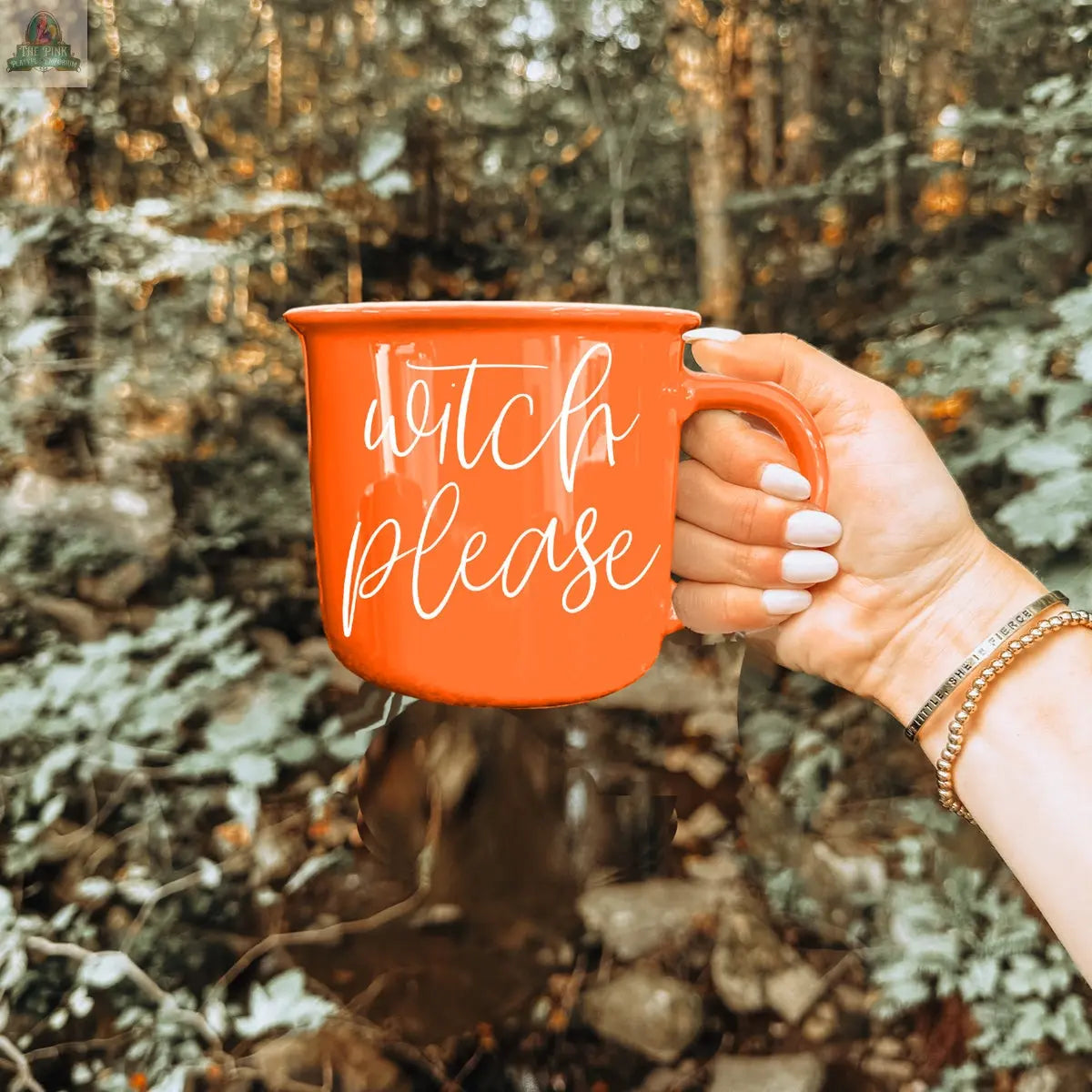 A hand with white-painted nails holds the Witch Please Mug, an orange ceramic cup with "witch please" in white script, set against a forest backdrop with green foliage and trees.