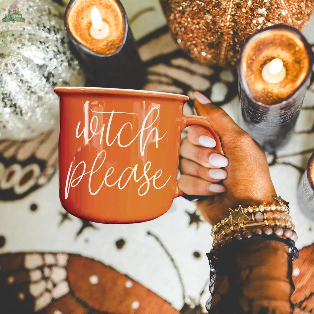 A hand with white-painted nails holds the Witch Please Mug, an orange ceramic cup surrounded by candles, glittery pumpkins, and autumn decor.