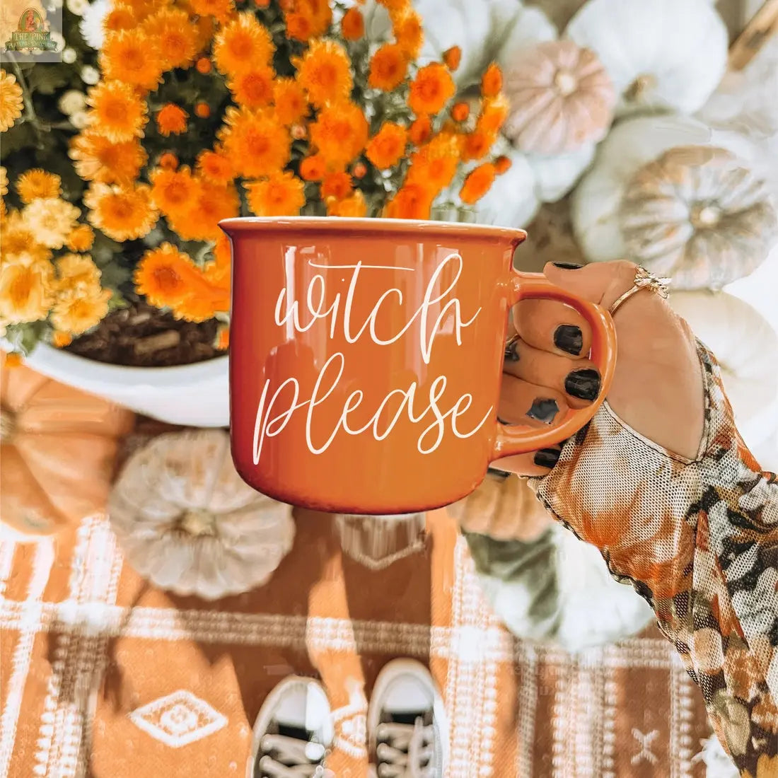 A hand with black-painted nails holds the Witch Please Mug. In the background, orange flowers and white pumpkins rest on a patterned orange rug, creating a festive Halloween vibe.
