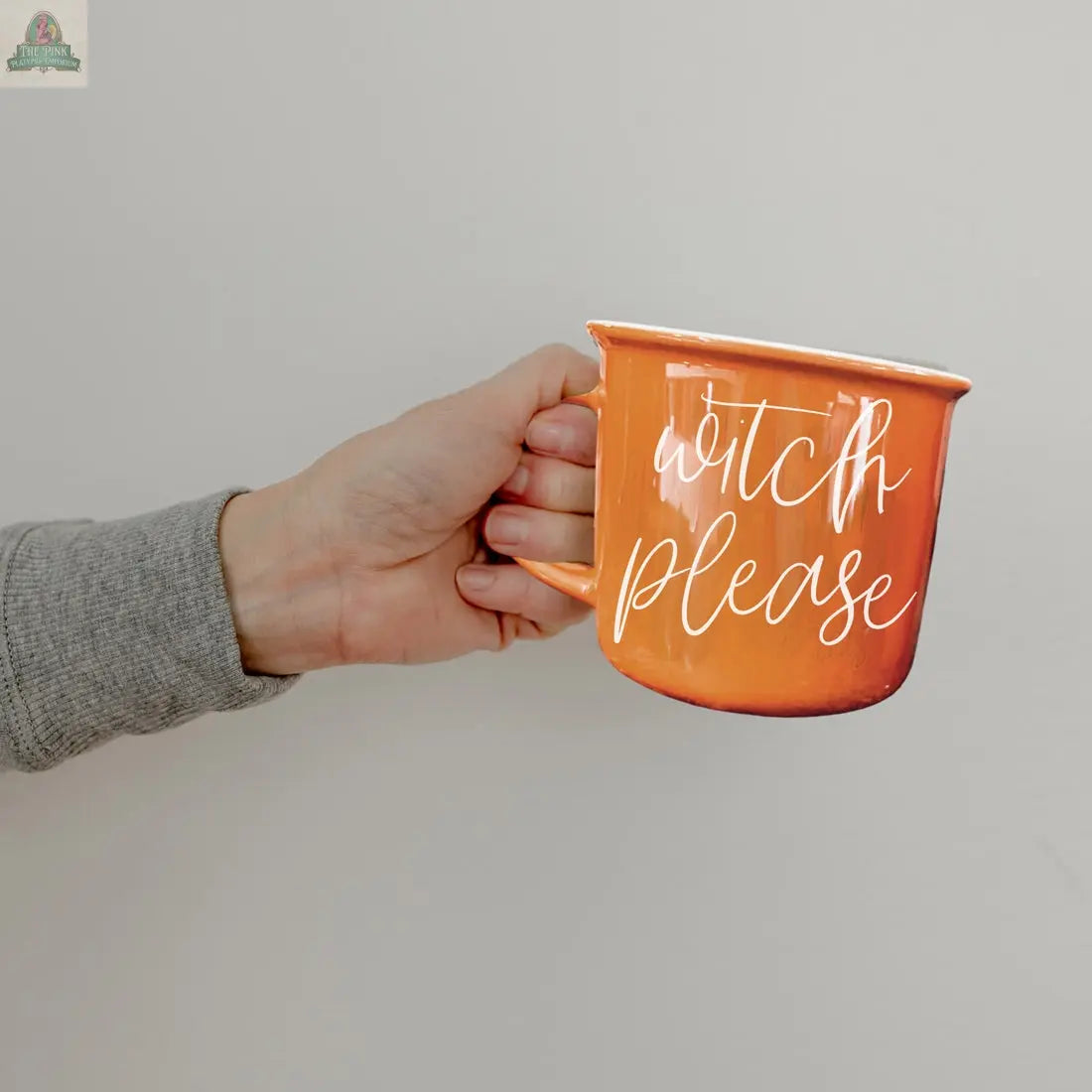 A hand holds the Witch Please Mug, an orange ceramic mug with "witch please" in white cursive lettering, set against a plain light background.