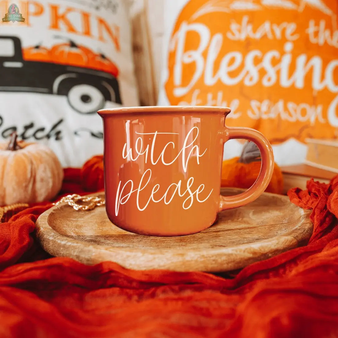 The Witch Please Mug, an orange ceramic mug with "Witch Please" in white script, sits on a wooden plate with autumn pillows, pumpkins, and cozy orange fabric in the background.
