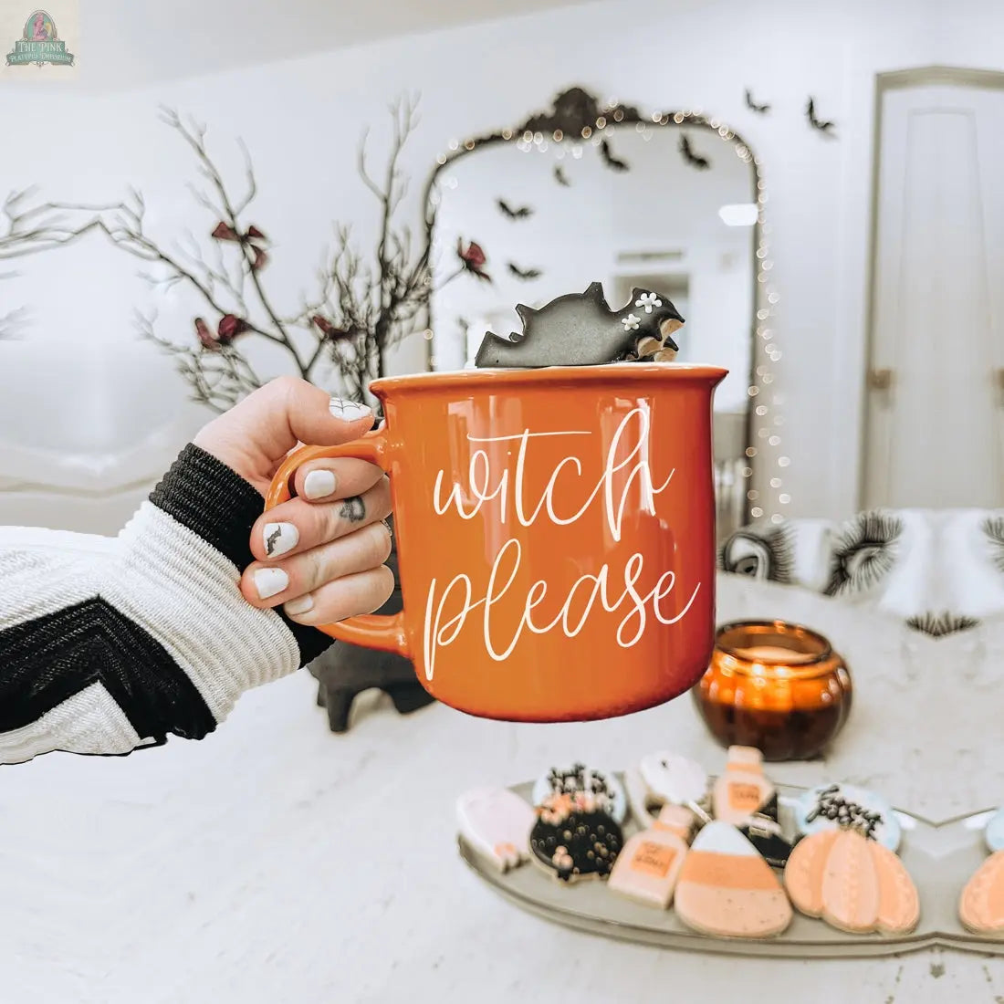 A hand with Halloween-themed nails holds the Witch Please Mug—a bright orange ceramic mug with a bat stirring stick inside. In the background are Halloween cookies, a lit candle, and festive seasonal decorations.