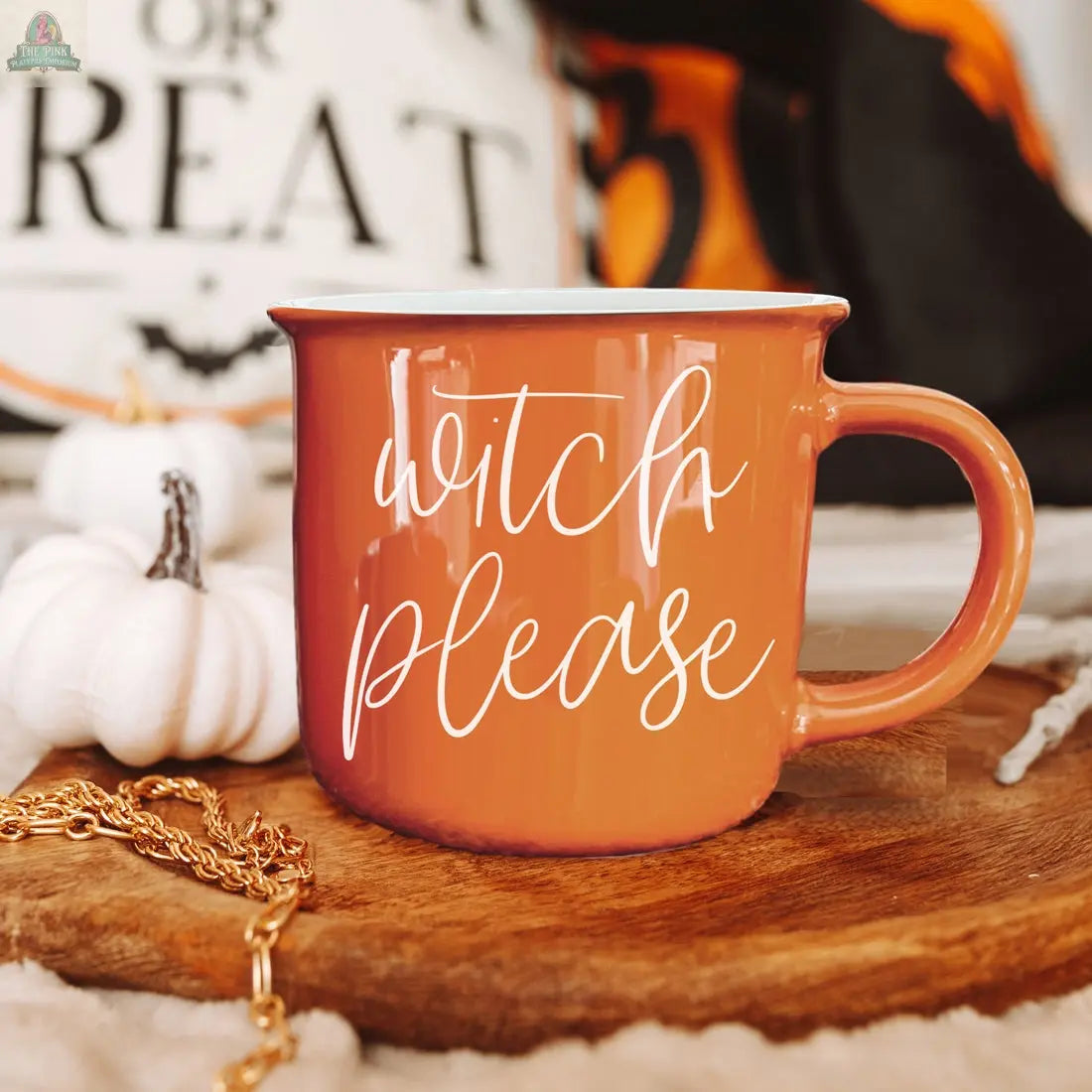 The Witch Please Mug, an orange ceramic mug with white script, rests on a wooden tray beside a white pumpkin and gold chain, surrounded by blurred Halloween decor.