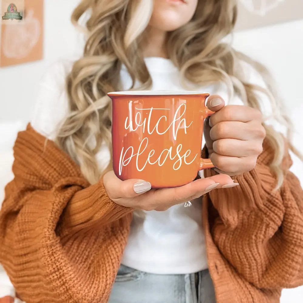 A woman with long wavy hair holds the Witch Please Mug, featuring “witch please” in white script. She wears a white shirt and a chunky burnt orange cardigan, with a softly blurred background adding a cozy vibe.