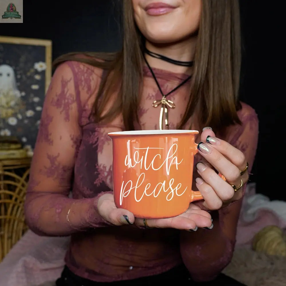 A woman in a sheer lacey top and choker holds the Witch Please Mug, a vibrant orange ceramic cup with white script. The cozy setting with owl art and spooky décor is perfect for enjoying your favorite drinks this Halloween season.