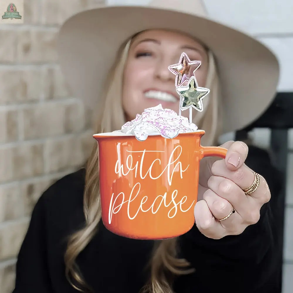 A smiling woman in a wide-brimmed hat holds the Witch Please Mug, topped with whipped cream and star-shaped stirrers, in front of a light brick wall.