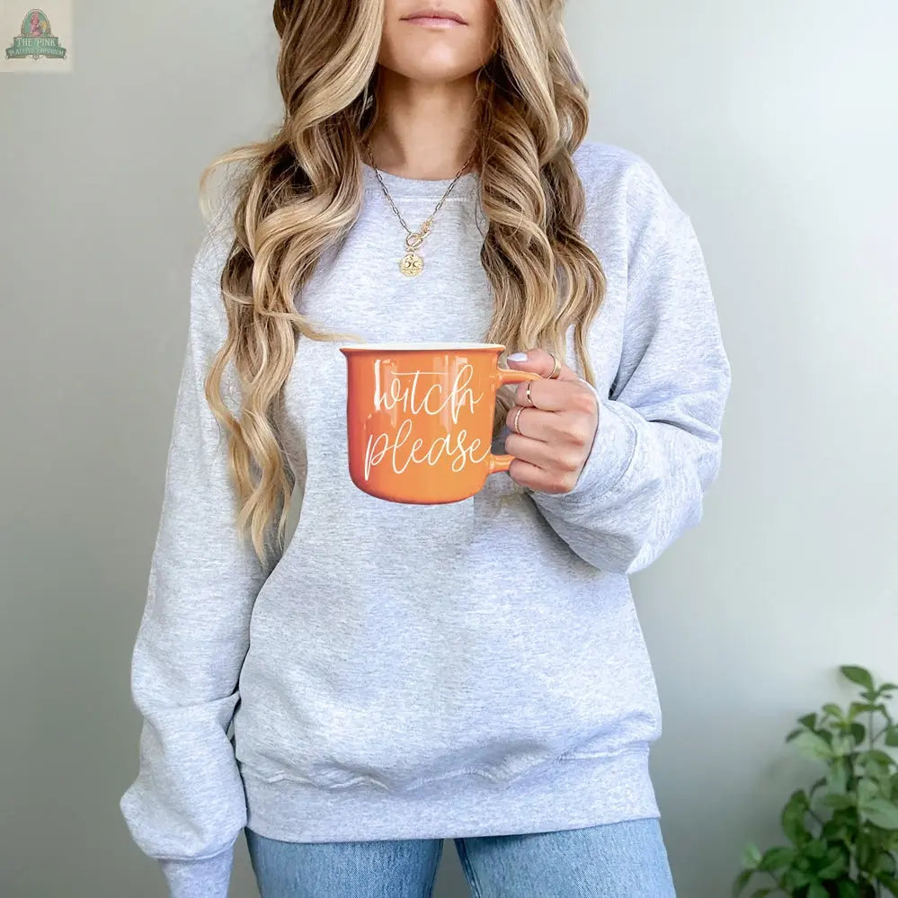 A woman with long wavy hair in a light gray sweatshirt and blue jeans holds the Witch Please Mug. Her face is partially out of frame as she stands against a plain background.
