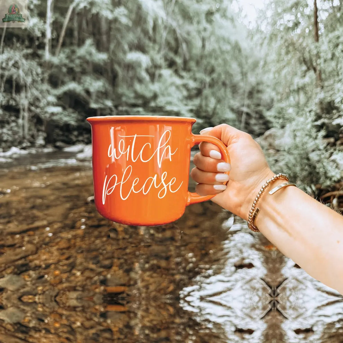 A hand wearing gold bracelets holds the Witch Please Mug, an orange ceramic cup with white script, in front of a forested creek—a bewitching Halloween coffee moment.