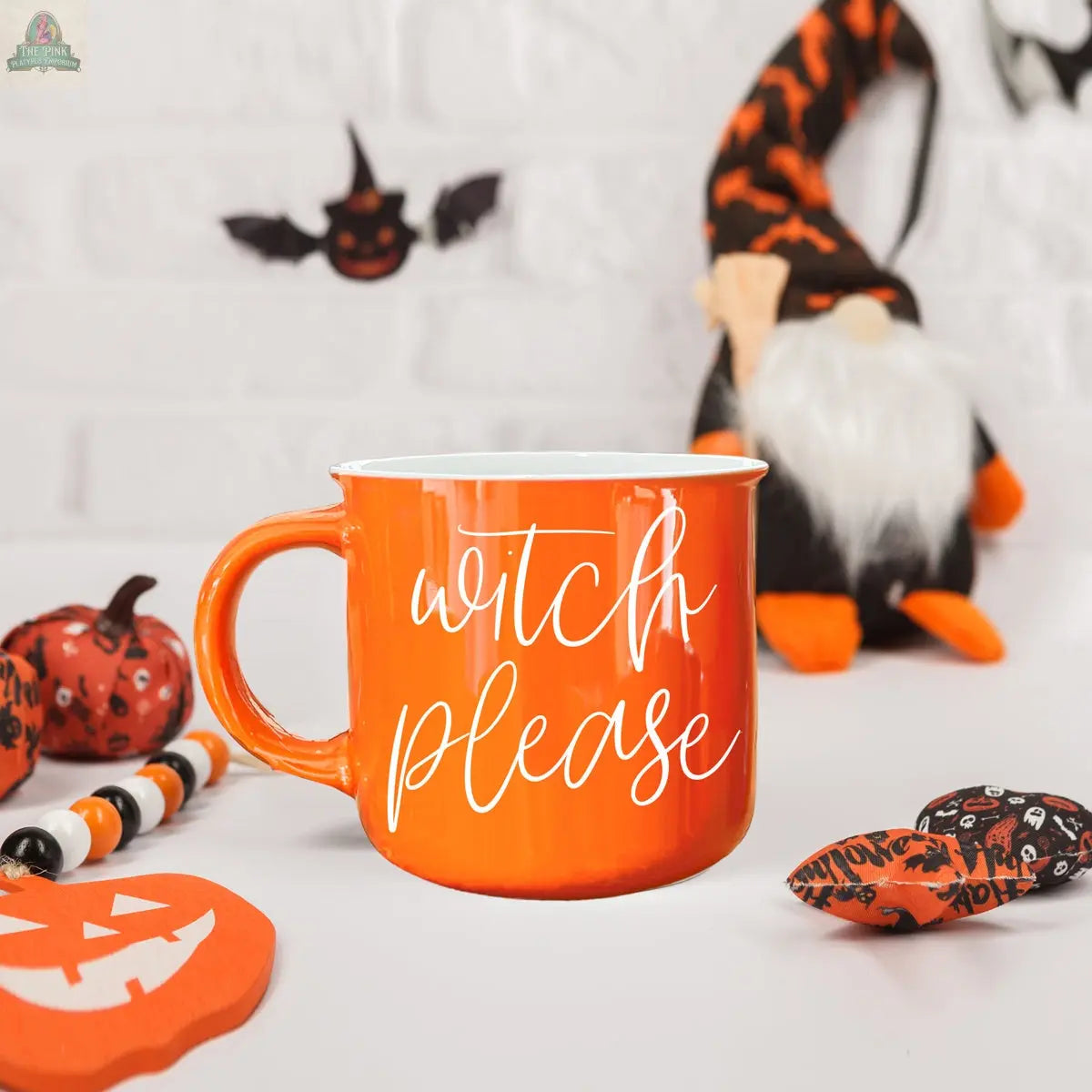 The Witch Please Mug, an orange ceramic mug with "witch please" text, rests on a white surface surrounded by Halloween décor—pumpkin, beads, gnome, and festive fabric—set against a white brick wall.