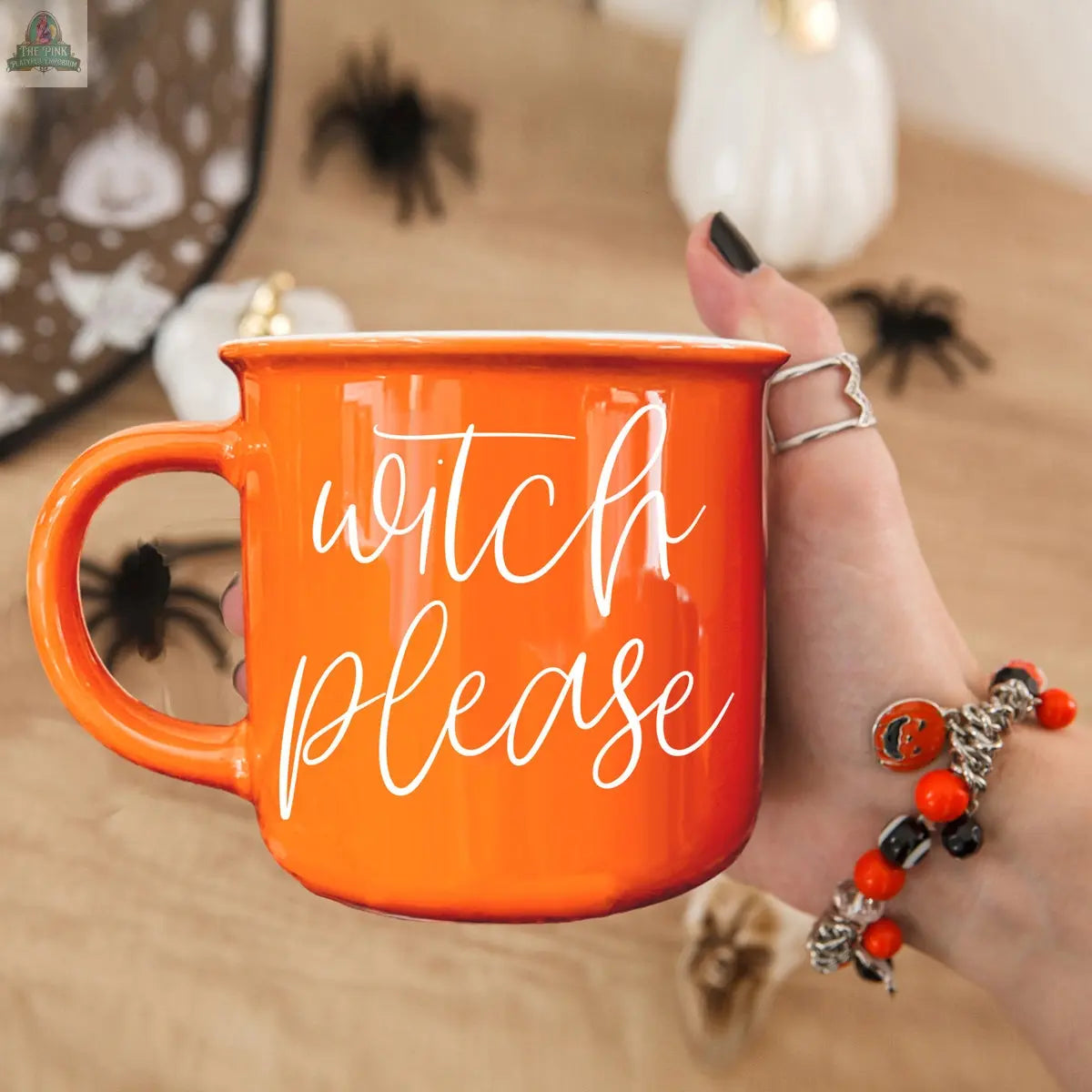 A hand with black nail polish holds the Witch Please Mug—an orange ceramic mug with white script. Halloween decorations like black spiders and a pumpkin are in the background, and the person wears orange and black festive jewelry.